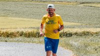 Ein Mann läuft auf einer Landstrasse. Er trägt ein weisses Cap, ein gelbes FRAGILE-T-Shirt und blaue kurze Hosen mit dem FRAGILE-Logo.