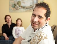 [Bitte in "Französich" uebersetzen:] Ein Mann dreht sich zur Kamera und lächelt. Im Hintergrund sind seine Frau und Tochter.