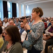 Eine Person am Rand des Publikums in einem Saal steht und hält ein Mikrofon in der Hand.