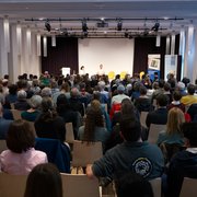 Sitzendes Publikum in einem Saal von hinten, mit Blick auf die Bühne. Es sind fast alle Stühle besetzt.