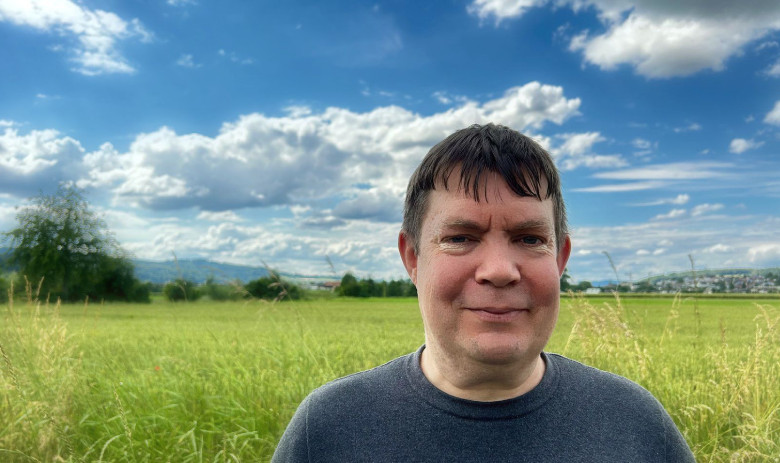 Un homme regarde la caméra. Il est dehors, dans la nature. En arrière-plan, une prairie verte et un ciel d'un bleu profond avec des nuages blancs.